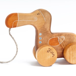Pu&ograve; includere: Un giocattolo di legno a traino a forma di dodo con un design dipinto in marrone, arancione e bianco. Il giocattolo ha una corda attaccata al becco e le parole "Finn" sono scritte su una delle ruote.