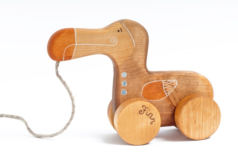 Pu&ograve; includere: Un giocattolo di legno a traino a forma di dodo con un design dipinto in marrone, arancione e bianco. Il giocattolo ha una corda attaccata al becco e le parole "Finn" sono scritte su una delle ruote.