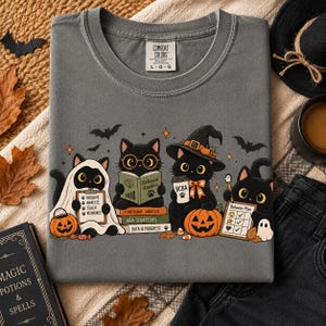 Puede incluir: Una camiseta gris carbón con un gráfico de cuatro gatos negros disfrazados para Halloween. Un gato lleva una sábana de fantasma, otro lee un libro titulado Behavior Analysis, otro lleva un sombrero de bruja y otro sostiene un plan de comportamiento.