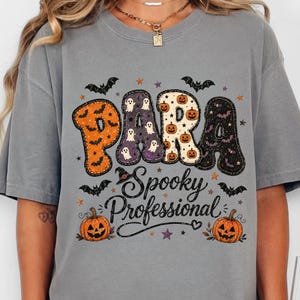May include: A grey short-sleeve t-shirt featuring a graphic print with the text "PARA Spooky Professional". The design includes orange pumpkins, purple ghosts, and black bats. The letters are filled with patterns of pumpkins, ghosts, and bats.