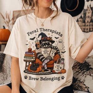 Op de afbeelding: Een crèmekleurig t-shirt met een afbeelding van een wasbeer met een heksenhoed en zonnebril, zittend in een stoel met een koffiebeker en klembord. De tekst luidt Feral Therapists en Brew Belonging, omringd door pompoenen, een spook en boeken.