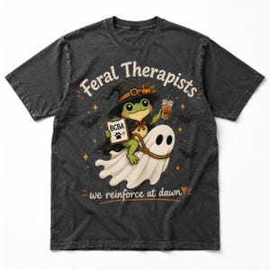 Könnte beinhalten: Ein anthrazitfarbenes T-Shirt mit einer Grafik eines grünen Frosches, der einen Hexenhut und einen Umhang trägt und auf einem weißen Geist reitet. Der Frosch hält ein Klemmbrett mit der Aufschrift BCBA, einem Pfotenabdruck und einem Kürbisgetränk. Text Feral Therapists we reinforce at dawn.