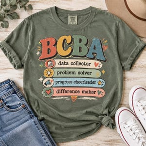 Op de afbeelding: Een olijfgroen T-shirt met korte mouwen en de tekst BCBA in grote, kleurrijke letters in retrostijl. Daaronder staan vier horizontale balken met de tekst data collector, problem solver, progress cheerleader en difference maker.