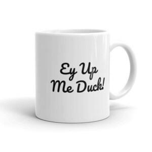 May include: White ceramic mug with a black script that reads "Ey Up Me Duck!". The mug has a curved handle and a glossy finish. The text is centered on the mug.