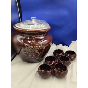 Op de afbeelding: Een vintage Heinz Oven Baked Beans keramische pot met een glazen deksel en zes bijpassende kleine bonenpotten. De grote pot is bruin met het Heinz-logo en de tekst. De deksel heeft een kleine handgreep.