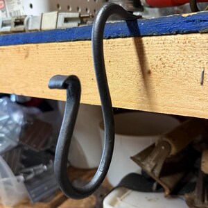 May include: A hand-forged, dark metal hook with a curved design. The hook is suspended from a wooden shelf, showcasing its unique shape and craftsmanship. The background includes tools and storage containers, suggesting a workshop setting.