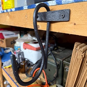 May include: A black, wrought iron hook mounted on a wooden shelf. The hook is curved and has a decorative design. The shelf is made of light-colored wood and is attached to a wall. The hook is designed for hanging items.