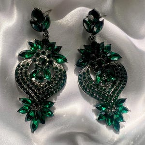 May include: A pair of ornate, emerald green earrings. Each earring features a cluster of green gemstones at the top, connected to a larger, heart-shaped design. The heart shape is filled with smaller green stones and a floral design at the bottom.
