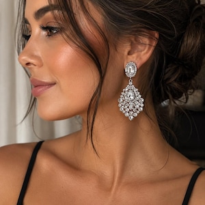 May include: Elegant silver-toned chandelier earrings with a sparkling, clear crystal design. The earrings feature a large, teardrop-shaped cluster of crystals, suspended from a smaller, rectangular crystal. A classic accessory for special occasions.