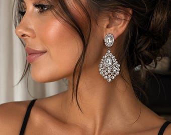 May include: Elegant silver-toned chandelier earrings with a sparkling, clear crystal design. The earrings feature a large, teardrop-shaped cluster of crystals, suspended from a smaller, rectangular crystal. A classic accessory for special occasions.