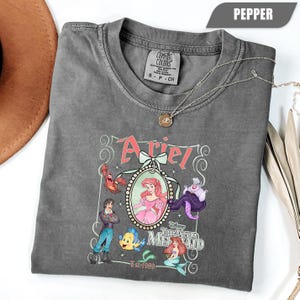 Puede incluir: Camiseta color pimienta con un dise&ntilde;o de Ariel de La Sirenita, con personajes como Sebasti&aacute;n, Flounder y &Uacute;rsula. El dise&ntilde;o incluye el nombre "Ariel" y el texto "The Little Mermaid Est-1989". Un collar plateado con un colgante est&aacute; sobre la camiseta.