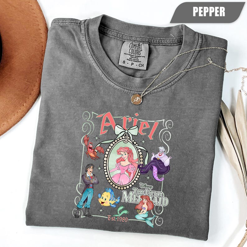 Puede incluir: Camiseta color pimienta con un dise&ntilde;o de Ariel de La Sirenita, con personajes como Sebasti&aacute;n, Flounder y &Uacute;rsula. El dise&ntilde;o incluye el nombre "Ariel" y el texto "The Little Mermaid Est-1989". Un collar plateado con un colgante est&aacute; sobre la camiseta.