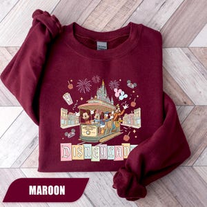 Pode incluir: Camisola bord&ocirc; com design da Disneyland. O gr&aacute;fico inclui um bonde com Mickey Mouse, Minnie, Donald e Pateta, juntamente com fogos de artif&iacute;cio e o logotipo da Disneyland. A camisola &eacute; de cor bord&ocirc; lisa.