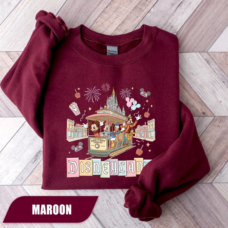 Pode incluir: Camisola bord&ocirc; com design da Disneyland. O gr&aacute;fico inclui um bonde com Mickey Mouse, Minnie, Donald e Pateta, juntamente com fogos de artif&iacute;cio e o logotipo da Disneyland. A camisola &eacute; de cor bord&ocirc; lisa.