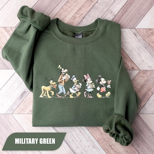 May include: Military green sweatshirt featuring a graphic of Disney characters Pluto, Goofy, Donald Duck, Minnie Mouse, and Mickey Mouse. The characters are walking and holding food items. The text "MILITARY GREEN" is at the bottom.