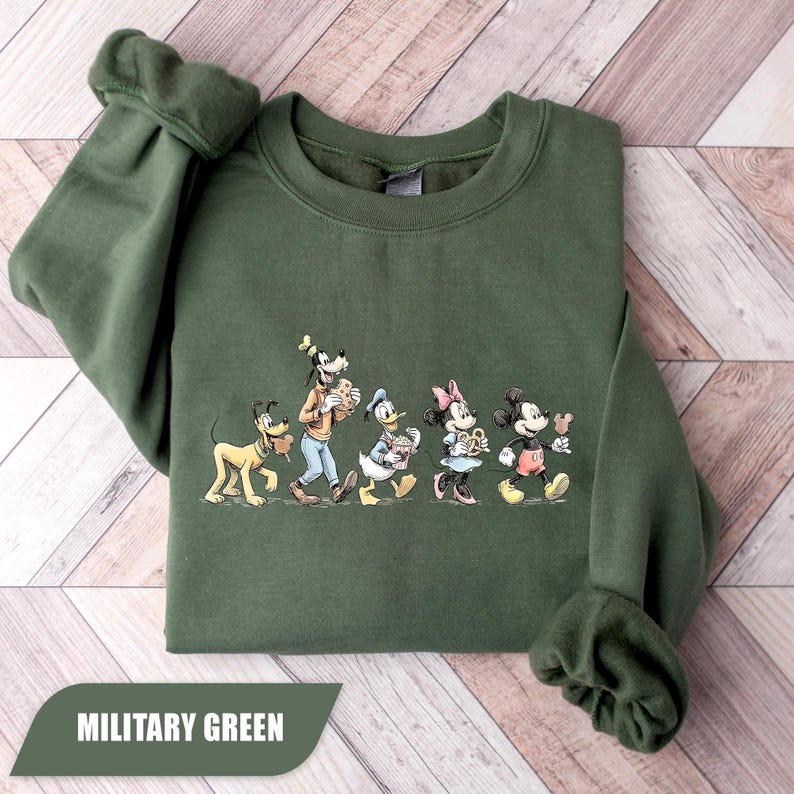 May include: Military green sweatshirt featuring a graphic of Disney characters Pluto, Goofy, Donald Duck, Minnie Mouse, and Mickey Mouse. The characters are walking and holding food items. The text "MILITARY GREEN" is at the bottom.