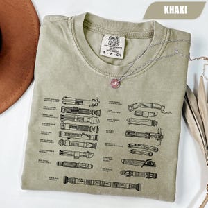 May include: Khaki-colored t-shirt with a graphic of various lightsabers and text. The shirt has a crew neck and short sleeves. A silver necklace with a pendant is draped over the shirt. The shirt is made by Comfort Colors.