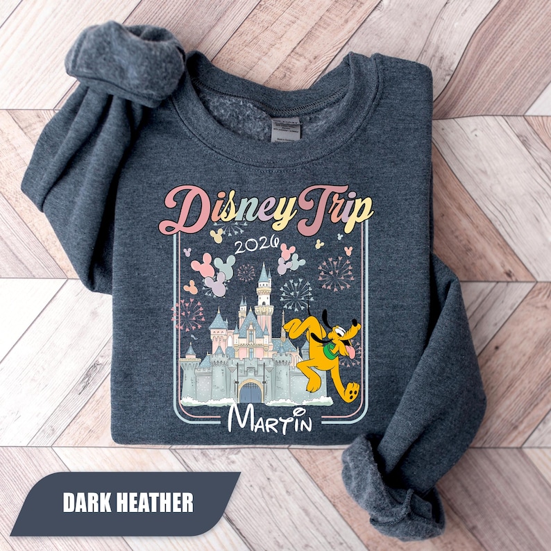 May include: Dark heather grey sweatshirt with a colourful graphic. The graphic features a castle, Pluto, and the words "Disney Trip 2026" with a personalized name. The sweatshirt is folded on a wooden surface.