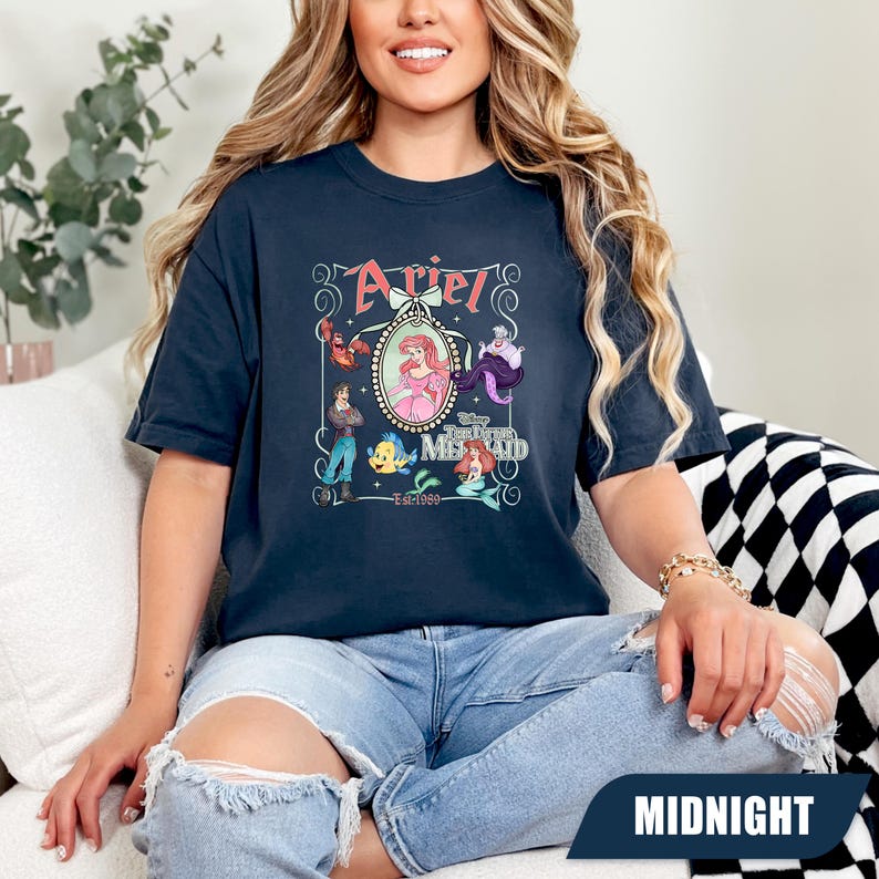 Puede incluir: Camiseta azul medianoche con un gr&aacute;fico de Ariel de La Sirenita. El dise&ntilde;o incluye la palabra "Ariel" en rosa, junto con ilustraciones de personajes de la pel&iacute;cula. Tambi&eacute;n se incluye el texto "The Little Mermaid" y "Est. 1989".