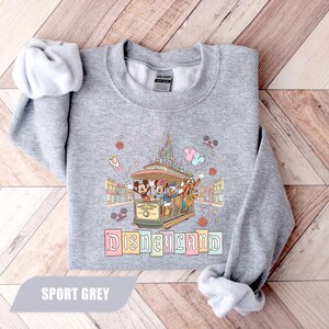 Pode incluir: Um moletom cinza claro com um design colorido da Disneyland. O gr&aacute;fico inclui Mickey Mouse, Minnie Mouse, Pato Donald e Pateta em um bonde, com a palavra "Disneyland" abaixo. O moletom &eacute; rotulado como "Sport Grey".