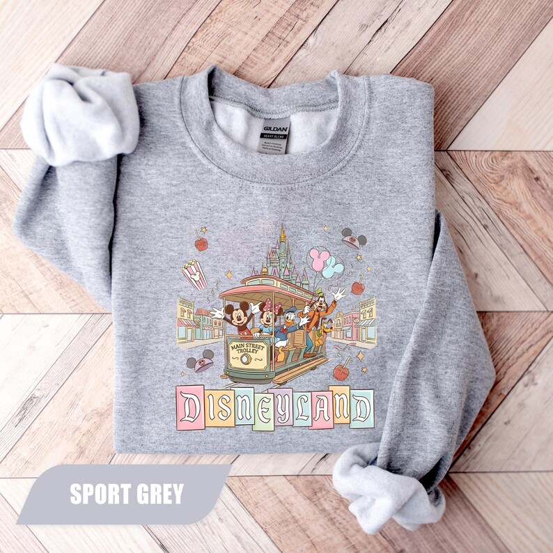 Pode incluir: Um moletom cinza claro com um design colorido da Disneyland. O gr&aacute;fico inclui Mickey Mouse, Minnie Mouse, Pato Donald e Pateta em um bonde, com a palavra "Disneyland" abaixo. O moletom &eacute; rotulado como "Sport Grey".