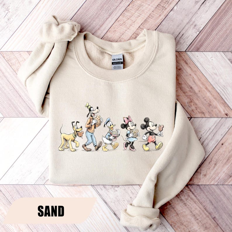 May include: Sand-colored sweatshirt featuring a graphic of Disney characters Pluto, Goofy, Donald Duck, Minnie Mouse, and Mickey Mouse. The characters are walking and holding treats. The word "SAND" is printed at the bottom of the sweatshirt.