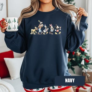 May include: Navy blue sweatshirt featuring a cartoon illustration of Disney characters including Goofy, Donald Duck, Minnie Mouse, and Mickey Mouse. The word "NAVY" is displayed on a rectangular label. A mug with Christmas tree designs is held in the hand.