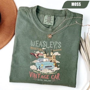 May include: A moss green t-shirt with a graphic featuring a vintage blue car, an owl, and a castle. The text reads "WEASLEY'S REGISTERED TRADEMARK" and "VINTAGE CAR". A silver necklace with a pendant is draped on the shirt.