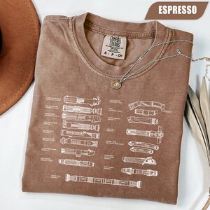 May include: Espresso-colored t-shirt with a diagram of lightsabers in white. The shirt has a crew neck and short sleeves. A silver necklace with a pendant is draped across the shirt. The shirt is labeled "Comfort Colors" and is size small.