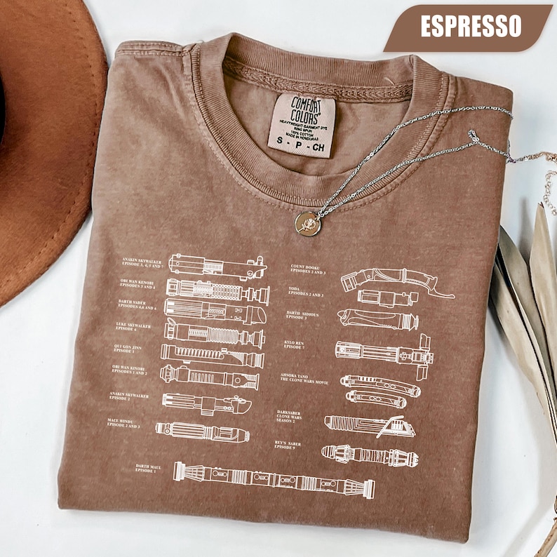 May include: Espresso-colored t-shirt with a diagram of lightsabers in white. The shirt has a crew neck and short sleeves. A silver necklace with a pendant is draped across the shirt. The shirt is labeled "Comfort Colors" and is size small.