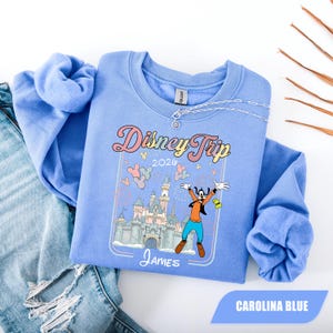 May include: Carolina blue sweatshirt featuring the text "Disney Trip 2020" and a graphic of Goofy in front of a castle. The name "James" is printed below the graphic. The sweatshirt is folded on top of a pair of blue jeans.