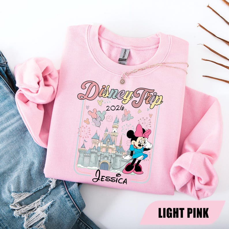 May include: Pale pink sweatshirt with a graphic of Minnie Mouse and a castle, with the text "Disney Trip 2020" and the name "Jessica". The sweatshirt has a crew neck and long sleeves.