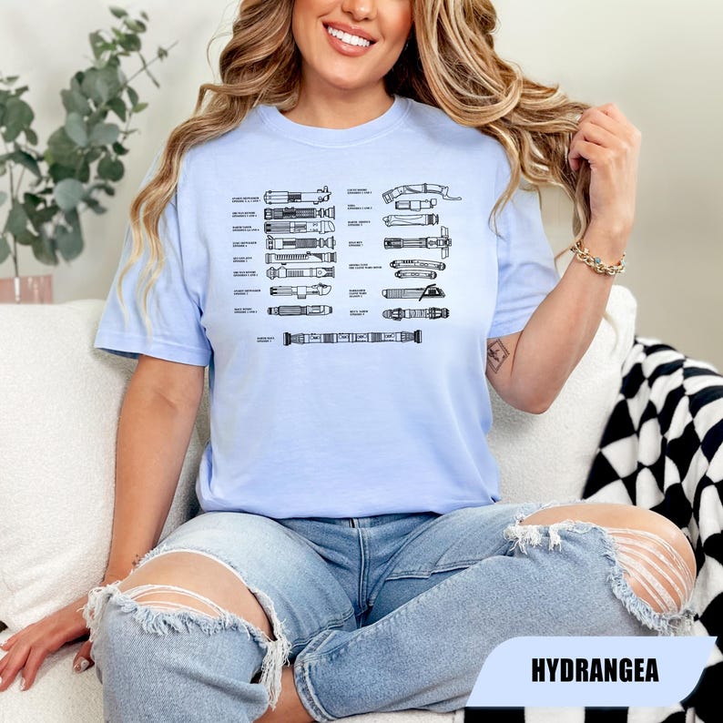 May include: A light blue t-shirt featuring a black line drawing of various lightsaber designs. The shirt is worn by a person sitting on a white couch. The word "HYDRANGEA" is printed on a white rectangle.