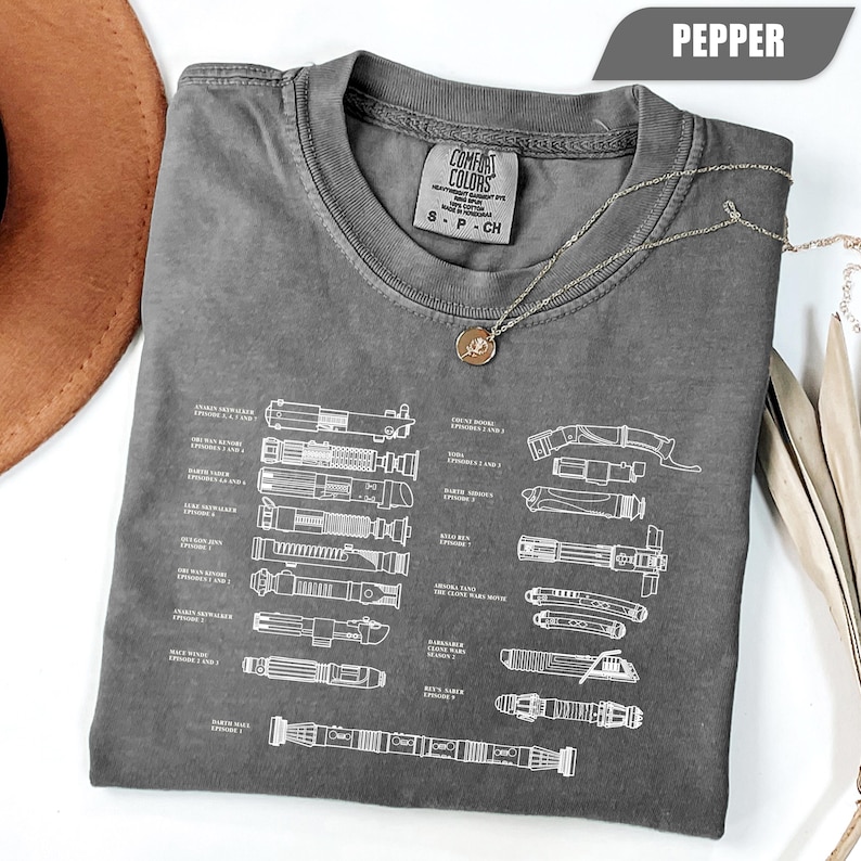 May include: A gray Comfort Colors t-shirt featuring a diagram of various lightsabers. The shirt has a crew neck and short sleeves. A silver necklace with a pendant is draped across the shirt. The shirt is labeled "Pepper" in the upper right corner.