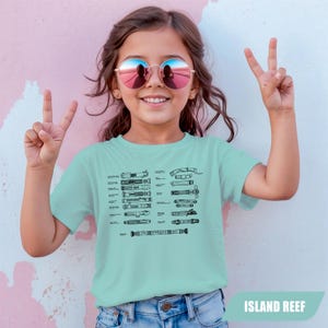 May include: A light blue t-shirt with a black diagram of various lightsabers. The shirt is worn by a young person wearing round sunglasses and making a peace sign with their fingers. The text "ISLAND REEF" is at the bottom right.