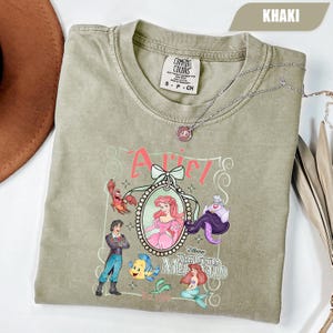 Puede incluir: Camiseta de color caqui con un dise&ntilde;o de Ariel de La Sirenita de Disney. El dise&ntilde;o incluye a Ariel, el Pr&iacute;ncipe Eric, Flounder, Sebasti&aacute;n y &Uacute;rsula. La camiseta tambi&eacute;n tiene el texto "Ariel" y "The Little Mermaid". Un collar plateado est&aacute; sobre la camiseta.