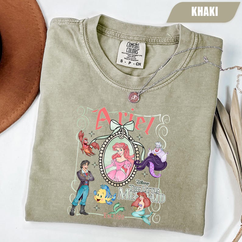 Puede incluir: Camiseta de color caqui con un dise&ntilde;o de Ariel de La Sirenita de Disney. El dise&ntilde;o incluye a Ariel, el Pr&iacute;ncipe Eric, Flounder, Sebasti&aacute;n y &Uacute;rsula. La camiseta tambi&eacute;n tiene el texto "Ariel" y "The Little Mermaid". Un collar plateado est&aacute; sobre la camiseta.