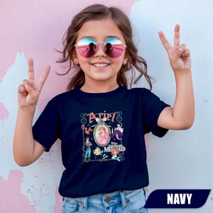 Puede incluir: Camiseta azul marino con un gr&aacute;fico colorido de Ariel de La Sirenita. El dise&ntilde;o incluye la palabra "Ariel" en una fuente decorativa, junto con otros personajes de la pel&iacute;cula. La camiseta es de estilo cuello redondo cl&aacute;sico.