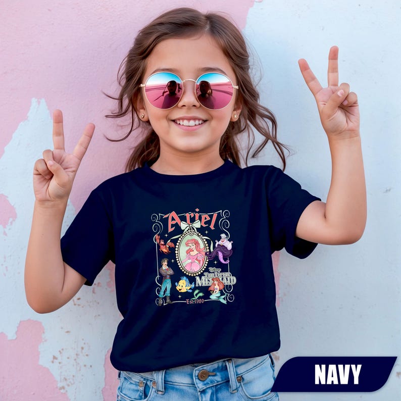 Puede incluir: Camiseta azul marino con un gr&aacute;fico colorido de Ariel de La Sirenita. El dise&ntilde;o incluye la palabra "Ariel" en una fuente decorativa, junto con otros personajes de la pel&iacute;cula. La camiseta es de estilo cuello redondo cl&aacute;sico.