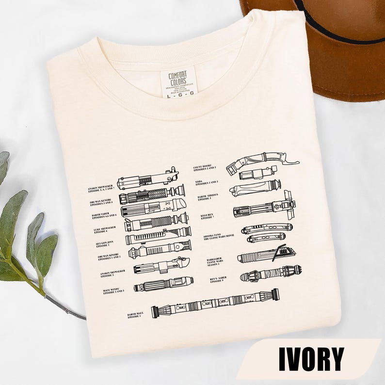 May include: Ivory-colored t-shirt featuring a detailed black line drawing of various lightsabers from the Star Wars saga. The shirt includes the text "IVORY" at the bottom and the Comfort Colors brand label.