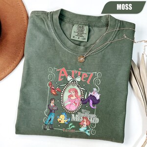 Puede incluir: Camiseta verde musgo con un gr&aacute;fico de Ariel de La Sirenita. El dise&ntilde;o incluye a Ariel, el Pr&iacute;ncipe Eric, Flounder, Sebasti&aacute;n y &Uacute;rsula. Tambi&eacute;n se muestra el texto "Ariel" y "The Little Mermaid".