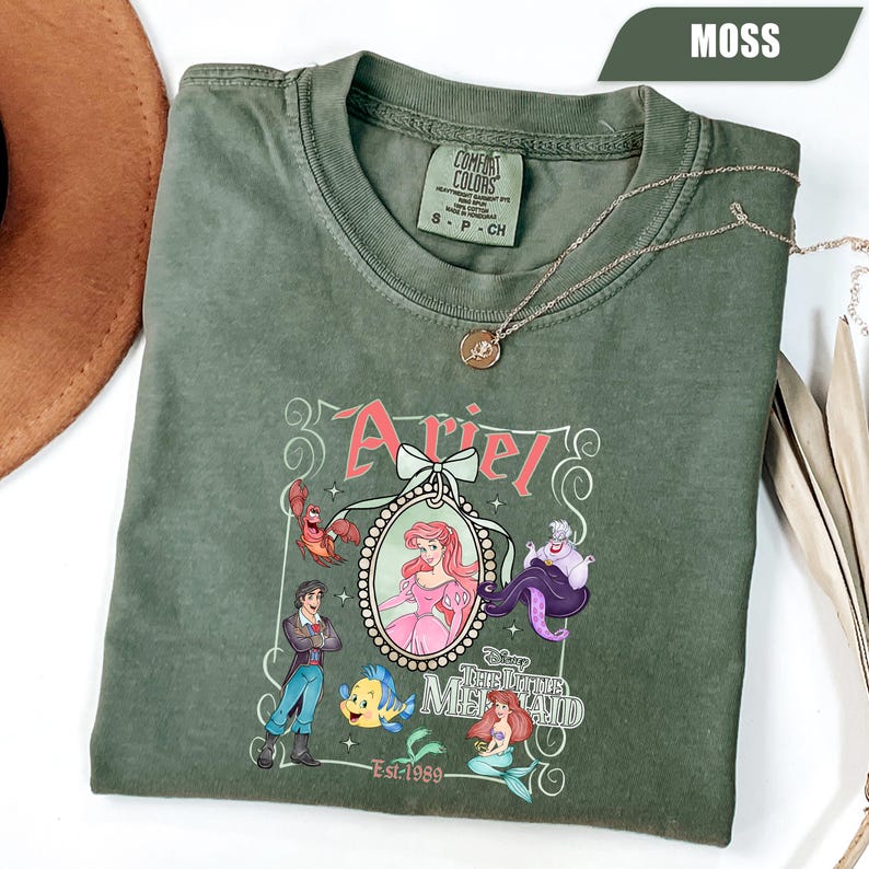 Puede incluir: Camiseta verde musgo con un gr&aacute;fico de Ariel de La Sirenita. El dise&ntilde;o incluye a Ariel, el Pr&iacute;ncipe Eric, Flounder, Sebasti&aacute;n y &Uacute;rsula. Tambi&eacute;n se muestra el texto "Ariel" y "The Little Mermaid".