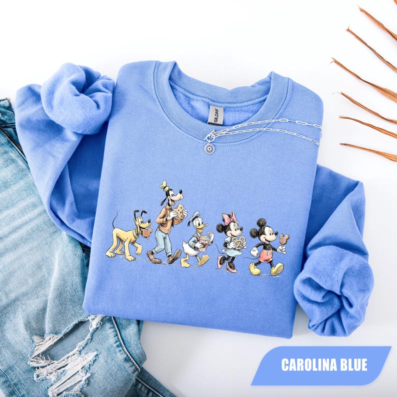 May include: Carolina blue sweatshirt featuring a graphic of Disney characters, including Mickey Mouse, Minnie Mouse, Donald Duck, Goofy, and Pluto. The characters are walking and holding snacks. The sweatshirt has a crew neck and long sleeves.