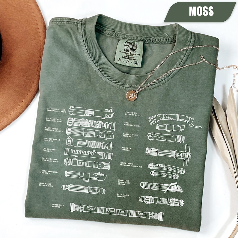 May include: A moss green t-shirt featuring a diagram of lightsabers with names and episode references. The shirt has a crew neck and short sleeves. A gold necklace with a pendant is draped over the shirt.