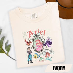 Puede incluir: Camiseta de color marfil con un dise&ntilde;o de Ariel de La Sirenita de Disney. El dise&ntilde;o incluye a Ariel, el Pr&iacute;ncipe Eric, Flounder, Sebasti&aacute;n y &Uacute;rsula. Tambi&eacute;n est&aacute;n presentes los textos "Ariel" y "The Little Mermaid".
