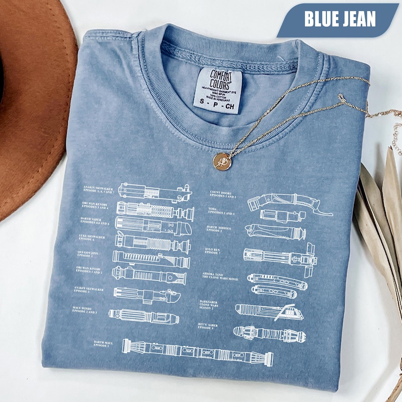 May include: Blue jean-colored t-shirt with a white schematic of lightsabers. The shirt has a crew neck and short sleeves. A gold necklace with a pendant is draped across the shirt. The tag reads "Comfort Colors".