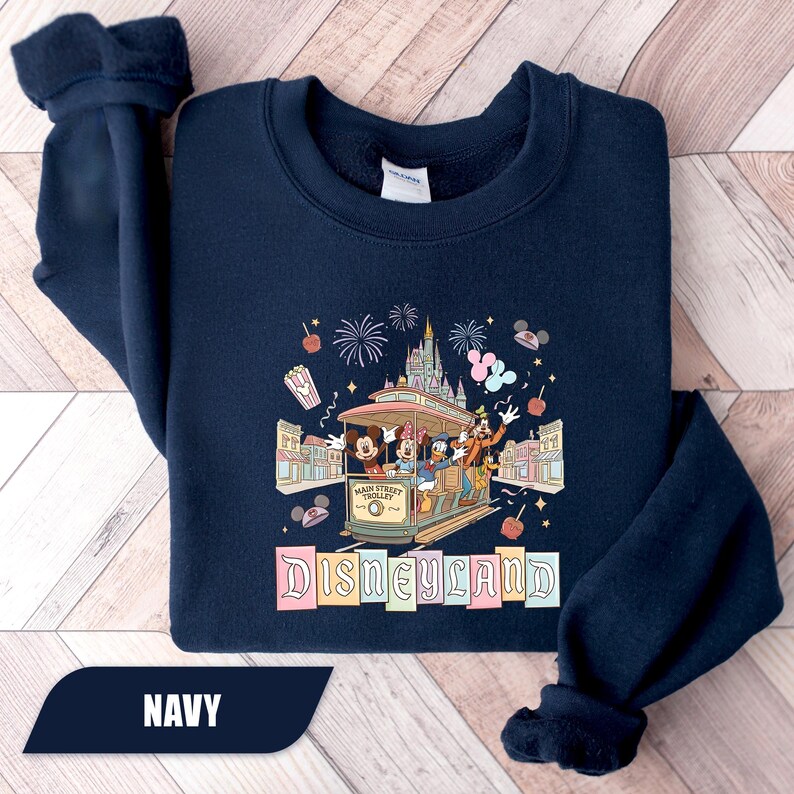 Pode incluir: Camisola azul marinho com um gr&aacute;fico colorido da Disneyland. O design inclui Mickey Mouse, Minnie, Pato Donald e Pateta num el&eacute;ctrico. A palavra "Disneyland" &eacute; exibida em cores pastel. Uma pe&ccedil;a de roupa confort&aacute;vel e casual.