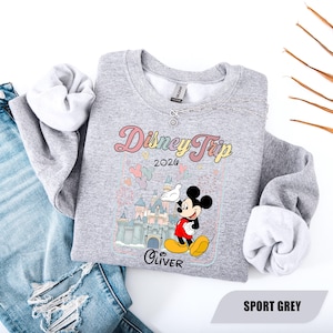 May include: Grey sweatshirt with a Disney Trip 2026 design. The design features Mickey Mouse, a castle, and the name "Oliver". The sweatshirt is paired with blue jeans.