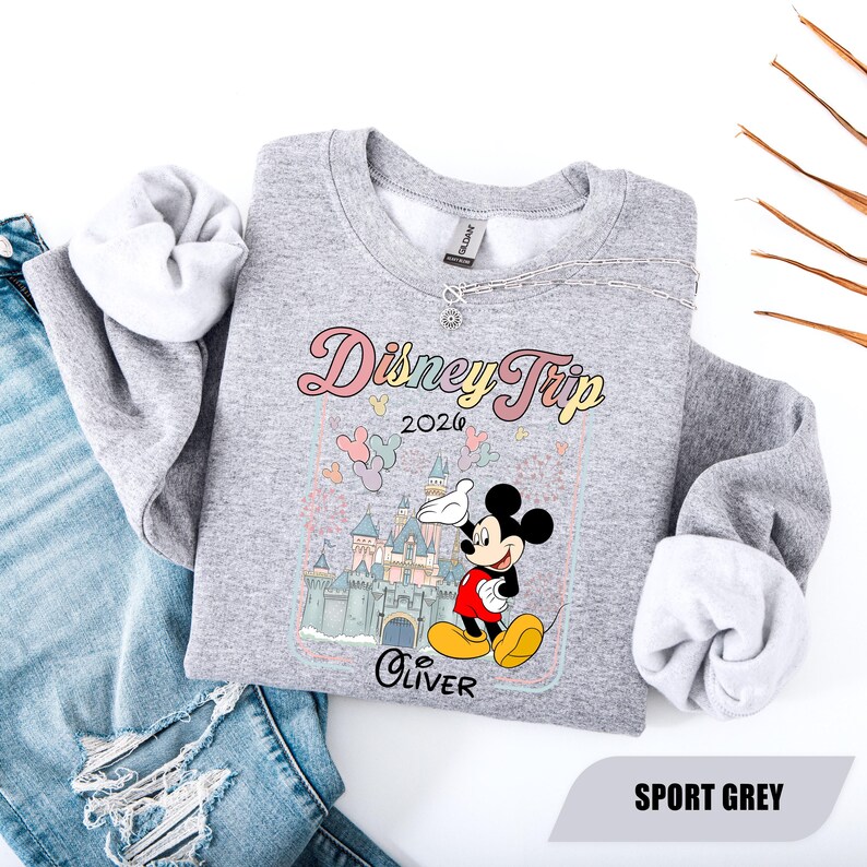 May include: Grey sweatshirt with a Disney Trip 2026 design. The design features Mickey Mouse, a castle, and the name "Oliver". The sweatshirt is paired with blue jeans.