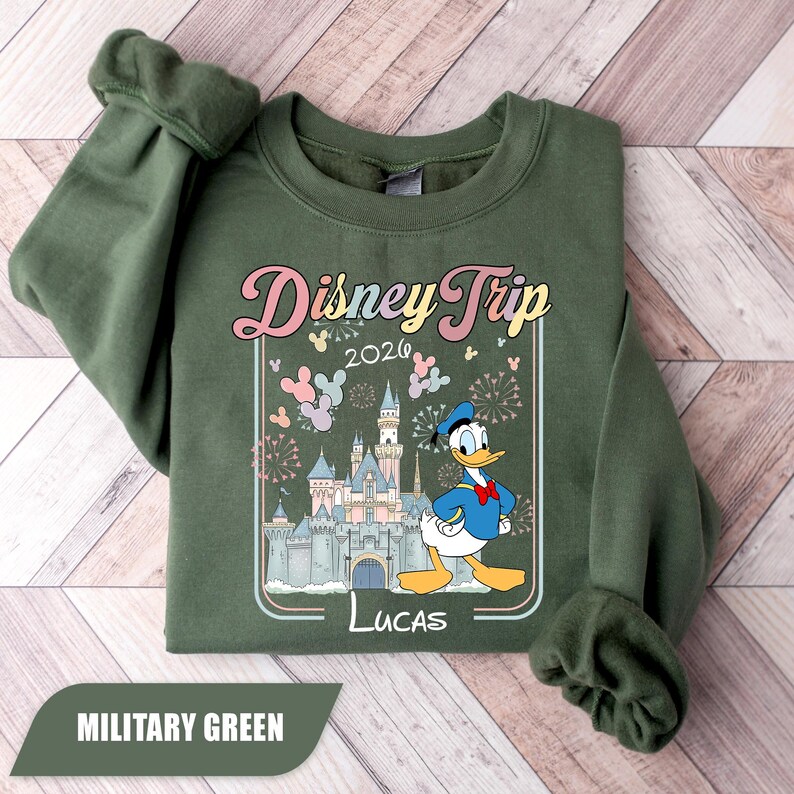May include: Military green sweatshirt with a Disney Trip 2026 design. The graphic includes Donald Duck, a castle, and the name "Lucas". The design is framed with a pastel border. A casual, comfortable style.